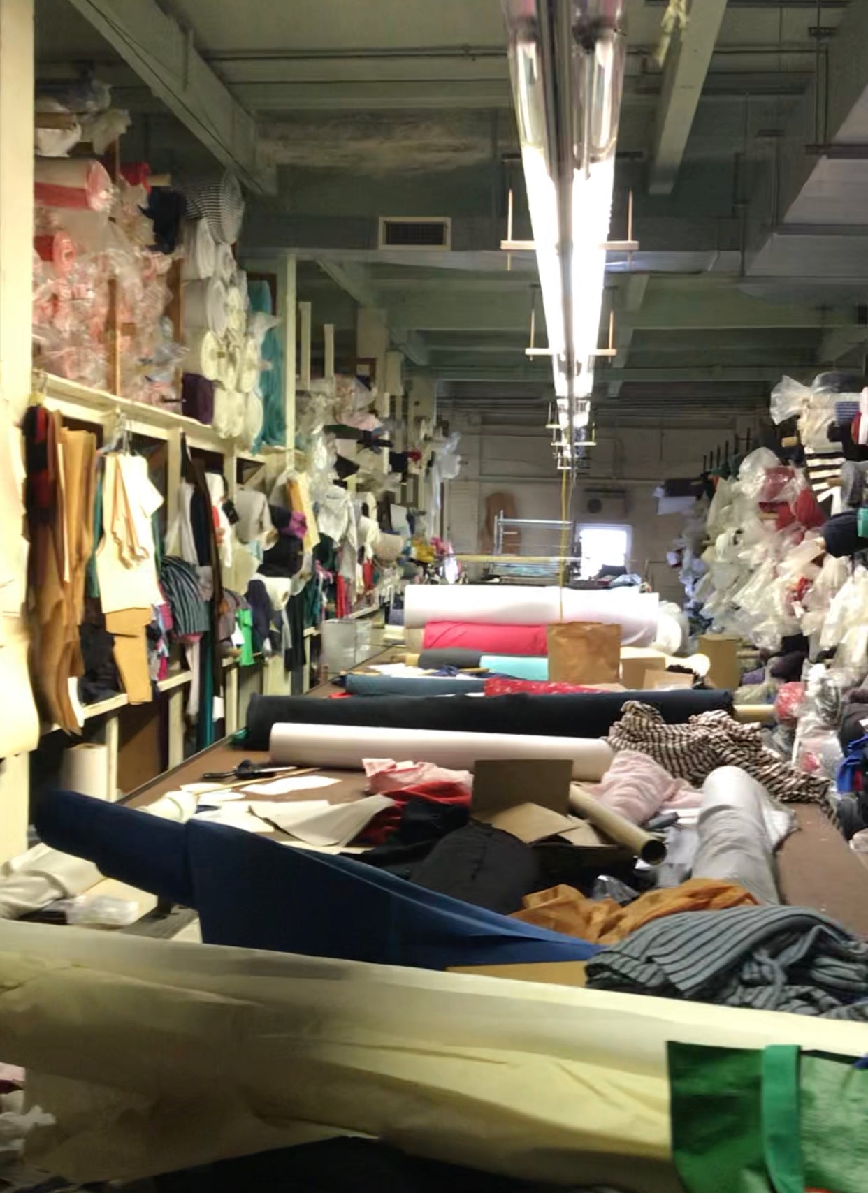 Old apparel warehouse with long work table covered in supplies and surrounded on both sides by floor to ceiling shelves of fabric rolls.